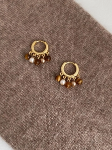 Détail de boucles d'oreilles créoles dorées avec oeil de tigre et perles sur tissu brun