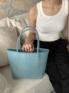 Close-up of a blue woven tote bag held by a model on a beige sofa in a lifestyle setting