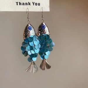 Macro detail of ethnic fish-shaped earrings with blue sequins and evil eye charm on white card