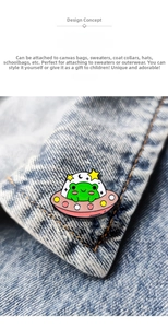 Close-up of a cartoon UFO frog enamel pin on blue denim fabric showing detailed craft