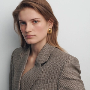 Close-up of a model wearing golden croissant style ribbed earrings with a brown blazer