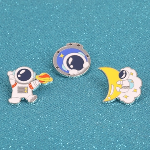Three astronaut enamel pins with moon and planet motifs on blue glitter background