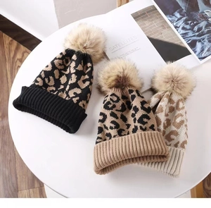 Close-up of leopard print knit beanies in black and brown with faux fur pom-poms on a table