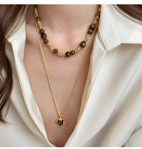Dettaglio di collana dorata a strati con perle occhio di tigre e ciondolo su camicia bianca