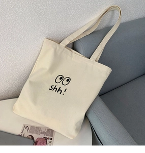 Detalle de bolso tote de lona beige con estampado de ojo sobre un sofá gris liso