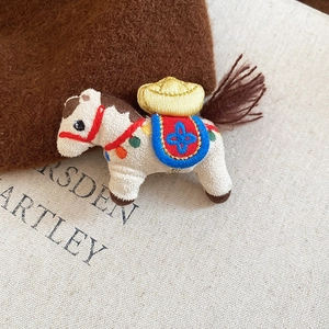 Close-up of a cream-white knit horse brooch with gold ingot embroidery and blue-red saddle