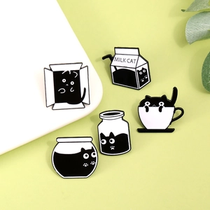 Close-up of five black cat enamel pins in various containers on a soft green surface