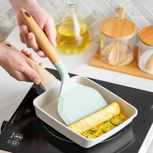Green silicone tamagoyaki spatula with wooden handle flipping an egg roll in a square pan