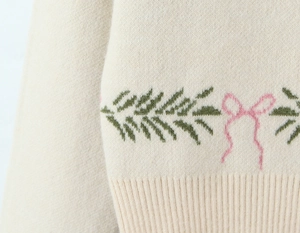 Detailed close-up of cream knit sweater with green pine leaf and pink bow embroidery