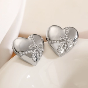 Silver heart stud earrings with sparkling rhinestone bow design on elegant ceramic surface
