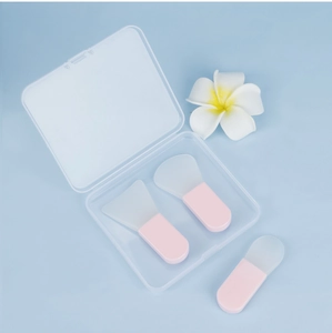 Three pink silicone face mask brushes with clear travel case on a blue floral background