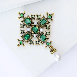 Baroque cross brooch with green stones and pearl pendant detail on a white pedestal
