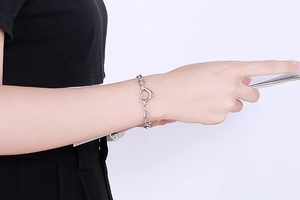 Detail of silver heart charm bracelet with OT clasp worn on wrist against white backdrop