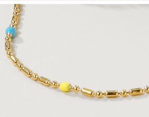 Makroaufnahme der Goldperlenkette mit blau-gelben Emaille-Akzenten auf cremeweißem Grund