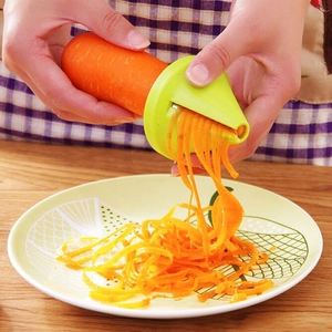 Green handheld vegetable spiralizer and grater slicing a fresh carrot on a wooden table