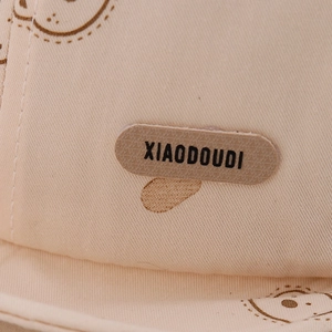 Detailed close-up of beige canvas baby hat with bear print and XIAODOUDI brand patch