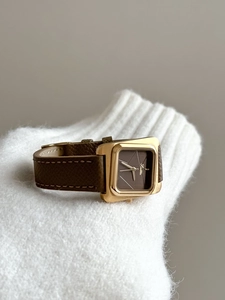 Gros plan d'une montre carrée dorée avec bracelet en cuir marron sur une manche blanche