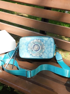 Blue crossbody bag with mandala patterns displayed on a wooden bench in natural sunlight