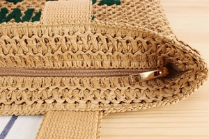 Detail view of woven straw tote bag featuring golden zipper and green embroidered pattern