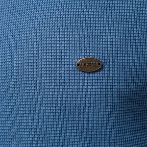 Détail de maille gaufrée bleue et plaque métallique AIOPESON sur chemise à manches longues