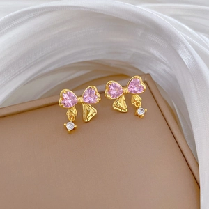 Dettaglio di orecchino a fiocco dorato con pietre a cuore rosa su portagioie beige