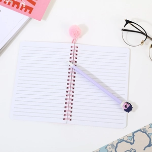 Pink spiral notebook set with a cartoon pen and fuzzy ball bookmark on a white desk