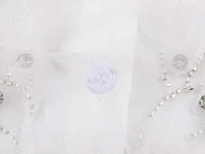 Détail d'une blouse blanche en dentelle montrant des perles argentées et un bouton blanc