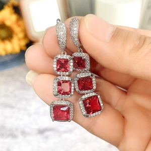 Close-up of dangling silver earrings featuring three square red stones with pavé diamonds