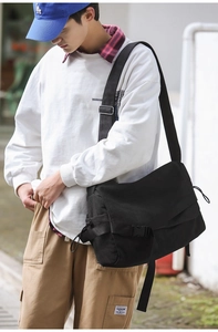 Detail of a black canvas messenger bag with buckle closure worn by a model in outdoor setting