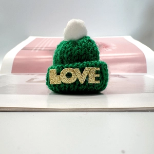 Detail einer grünen Mini-Strickmütze mit weißem Bommel und goldenem LOVE-Schriftzug