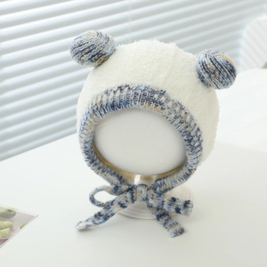 Close-up of white plush baby winter hat with double ear pom-poms and blue braided straps