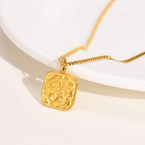 Close-up of a gold square embossed pendant on a fine box chain over a white ceramic plate