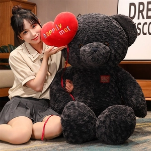 Detail of large black teddy bear plush with red heart pillow shown in home setting