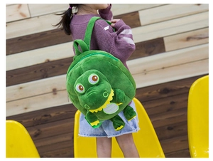 Detail view of a green plush crocodile backpack worn by a toddler against wooden wall
