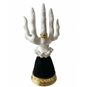 Detailed view of a white hand-shaped jewelry holder featuring a golden skull ring on velvet base