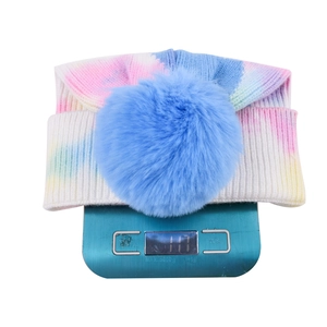 Tie-dye knit beanie with blue faux fur pom pom being weighed at 117g on a scale