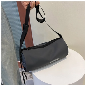 Détail d'un sac cylindrique gris foncé avec une large sangle noire sur socle blanc