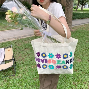 White canvas tote bag with floral embroidery and top handles shown in an outdoor park setting