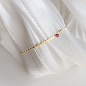 Detail of gold bead bracelet with polished bar and red heart pendant on white fabric
