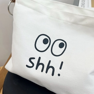 Close-up of white canvas tote bag featuring black eye graphics and shh text detail