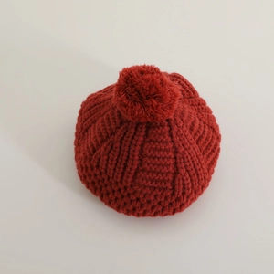 Détail du béret en maille rouge avec un grand pompon sur fond gris clair