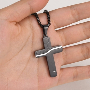 Detail of hand holding a black stainless steel cross pendant with white wave patterns and a diamond