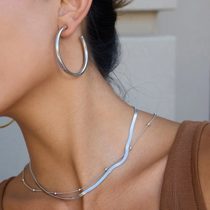 Detail view of large silver C-hoop earrings styled with layered snake bone chain necklaces