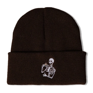 Dark brown knitted beanie with white skull embroidery holding coffee cup on a plain background