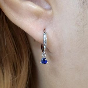 Close-up of silver earrings featuring a round deep blue sapphire pendant on human ear