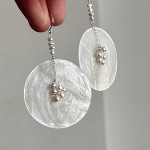 Hand holding elegant shell disc earrings with pearl drop tassel on silver hooks