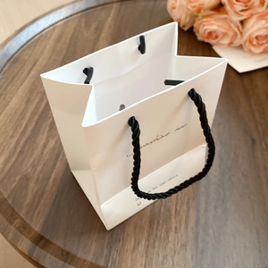Elegant white paper gift bag with black handles on a wooden table with pink roses background