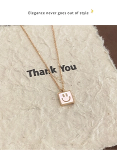 Close-up of golden necklace featuring a white square smiley face pendant on a textured card