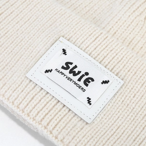 Detail of cream rib-knit beanie showing the embroidered SWIE logo patch and woven texture