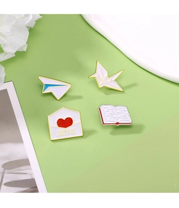 Detail of four enamel pins including paper plane, crane, book, and love letter on a green desk
